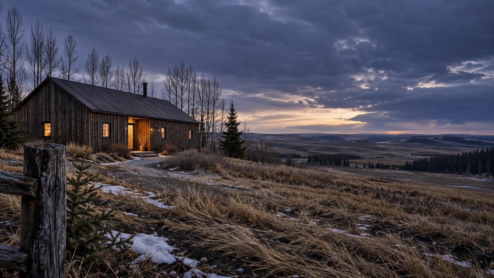 Imagined weathered timber building with a restrained warm doorway, close fence post, thawing snow and tawny grass overlooking a broad rolling prairie valley at winter dusk