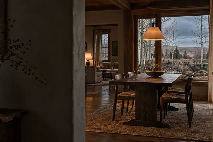 Imagined shadowed country interior with a plaster doorway, dark oak dining table, quiet living room, warm lamps and a timber window onto bare aspen, a kitchen garden and prairie ridges