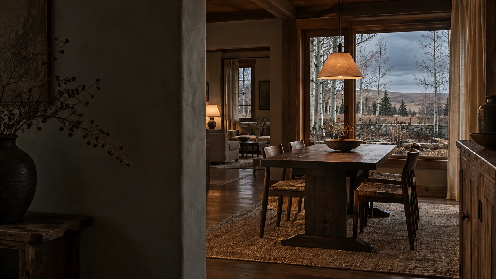 Imagined shadowed country interior with a plaster doorway, dark oak dining table, quiet living room, warm lamps and a timber window onto bare aspen, a kitchen garden and prairie ridges