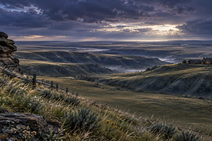 Imagined southern Alberta prairie at dusk, with close sandstone and grasses, overlapping coulee ridges and a small country home beneath a silver horizon.